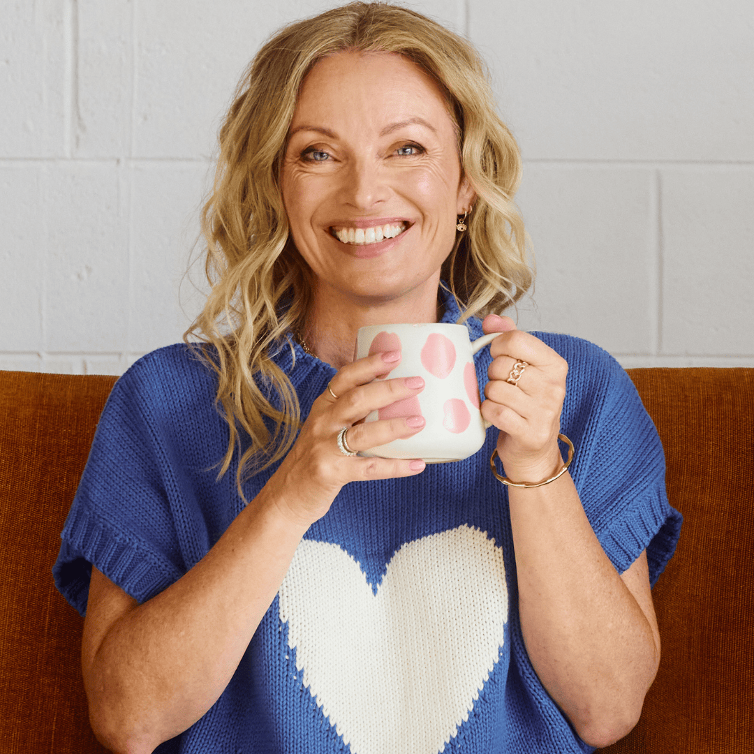 Lisa Walker, co-founder of Eir Women, smiling warmly while holding a pink polka dot mug, wearing a blue sweater with a white heart, seated on a rust-colored couch.