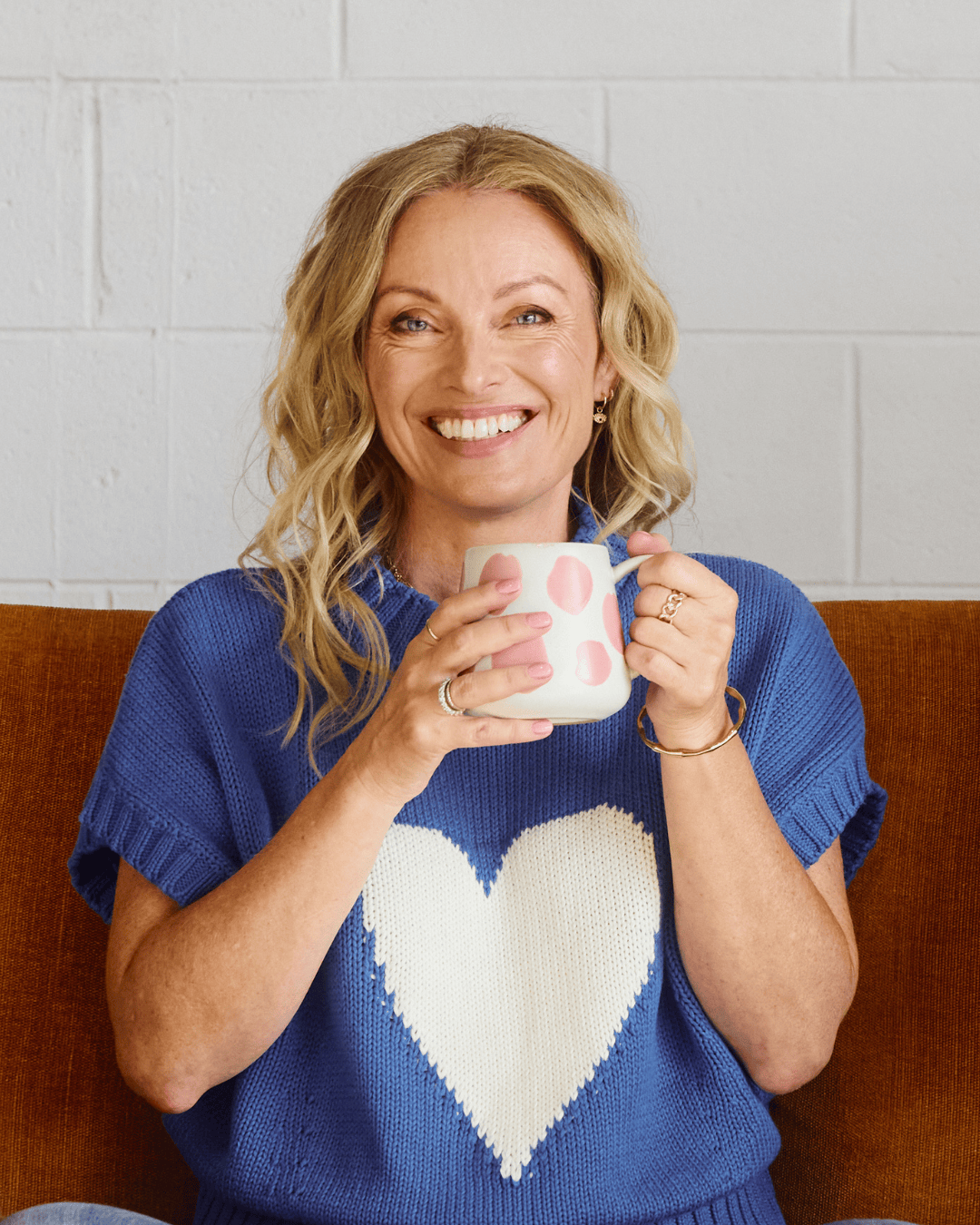 Lisa Walker, co-founder of Eir Women, smiling warmly while holding a pink polka dot mug, wearing a blue sweater with a white heart, seated on a rust-colored couch.