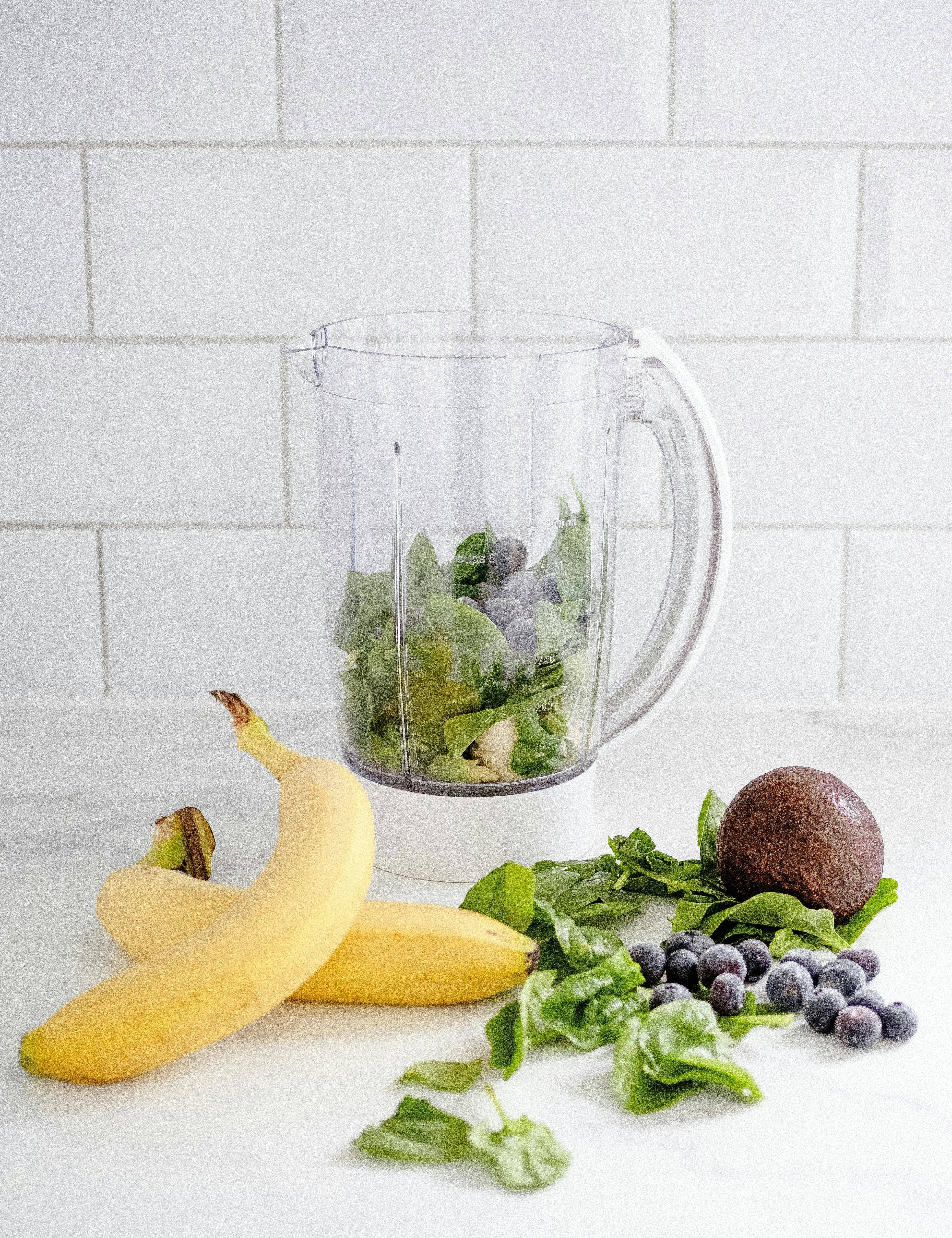 Healthy breakfast to balance blood sugar. Ingredients for a green smoothie, including bananas, spinach, avocado, and blueberries, arranged next to a blender jar on a white kitchen counter with a tiled backsplash.