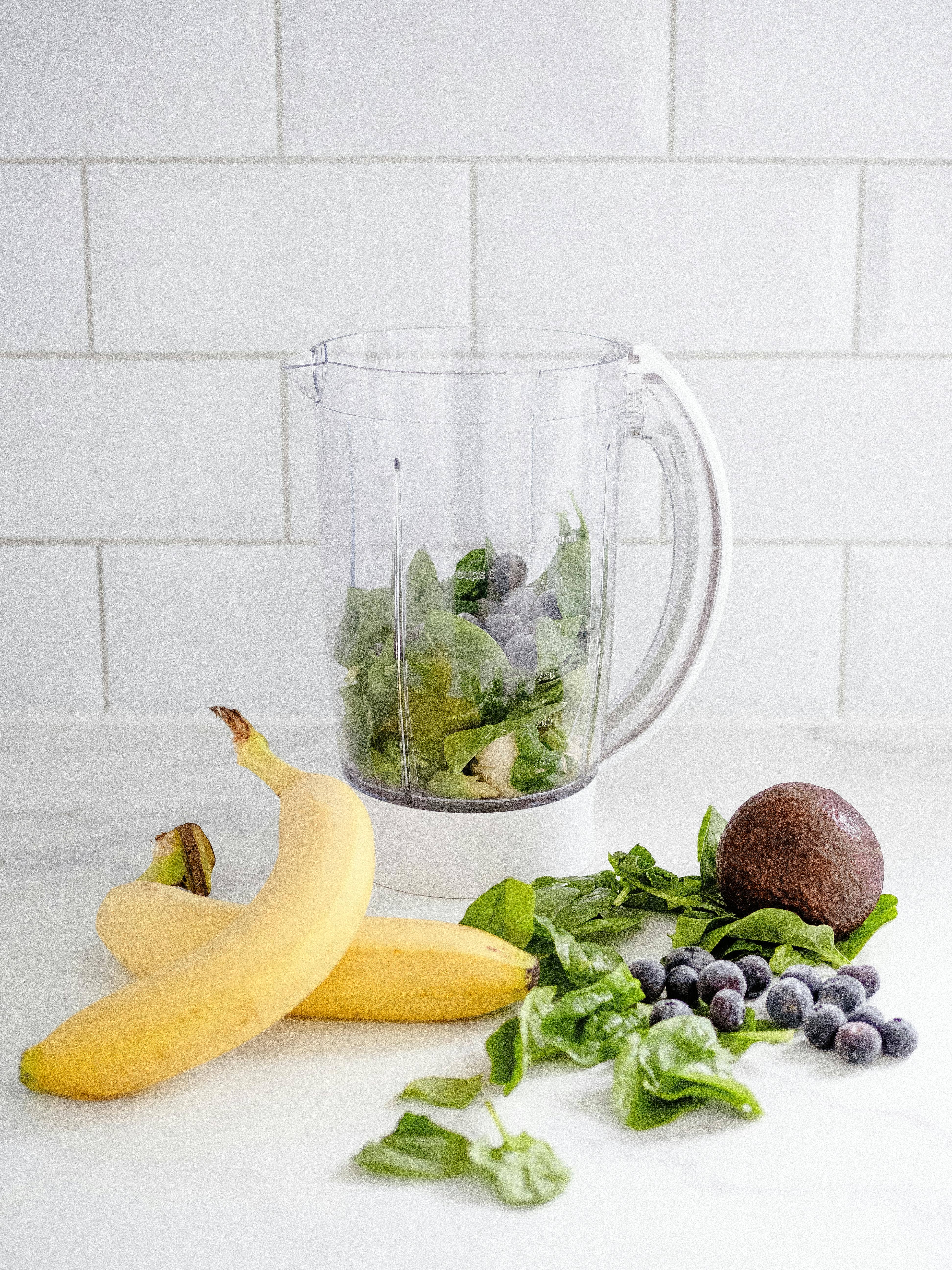 Healthy breakfast to balance blood sugar. Ingredients for a green smoothie, including bananas, spinach, avocado, and blueberries, arranged next to a blender jar on a white kitchen counter with a tiled backsplash.