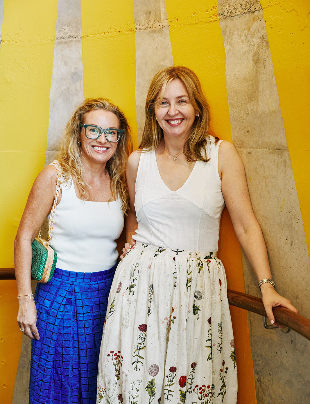two women with blonde hair standing together as friends. Both in white tops and coloured skirts.
