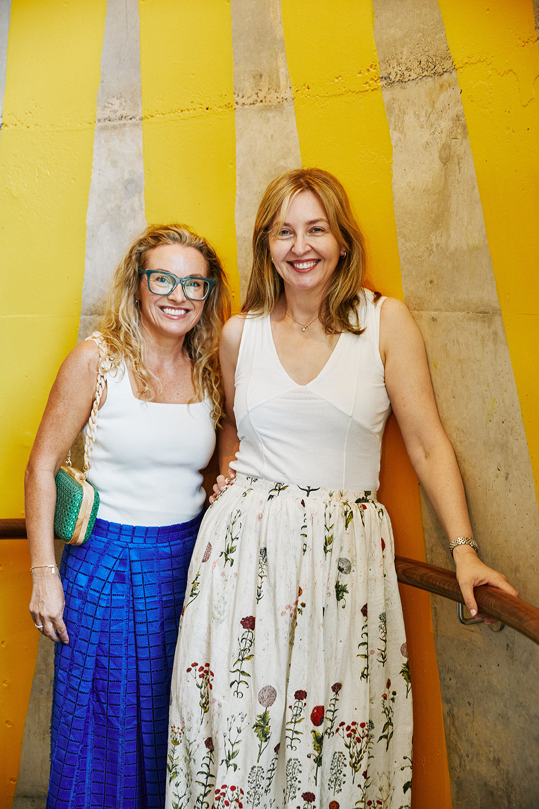 two women with blonde hair standing together as friends. Both in white tops and coloured skirts.