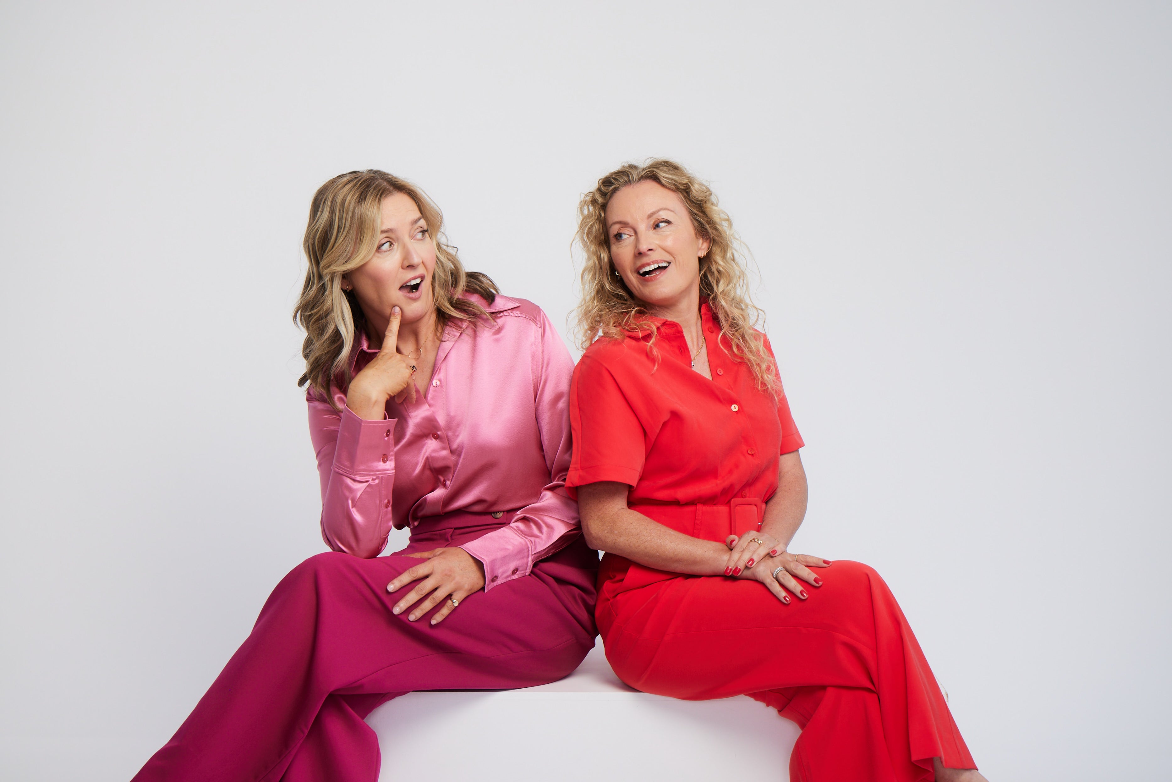 two women sitting together on white seat. One is wearing red jumpsuit and has long blonde curly hair and the other is wearing a pink suit. Both looking at each other and pulling a face. 