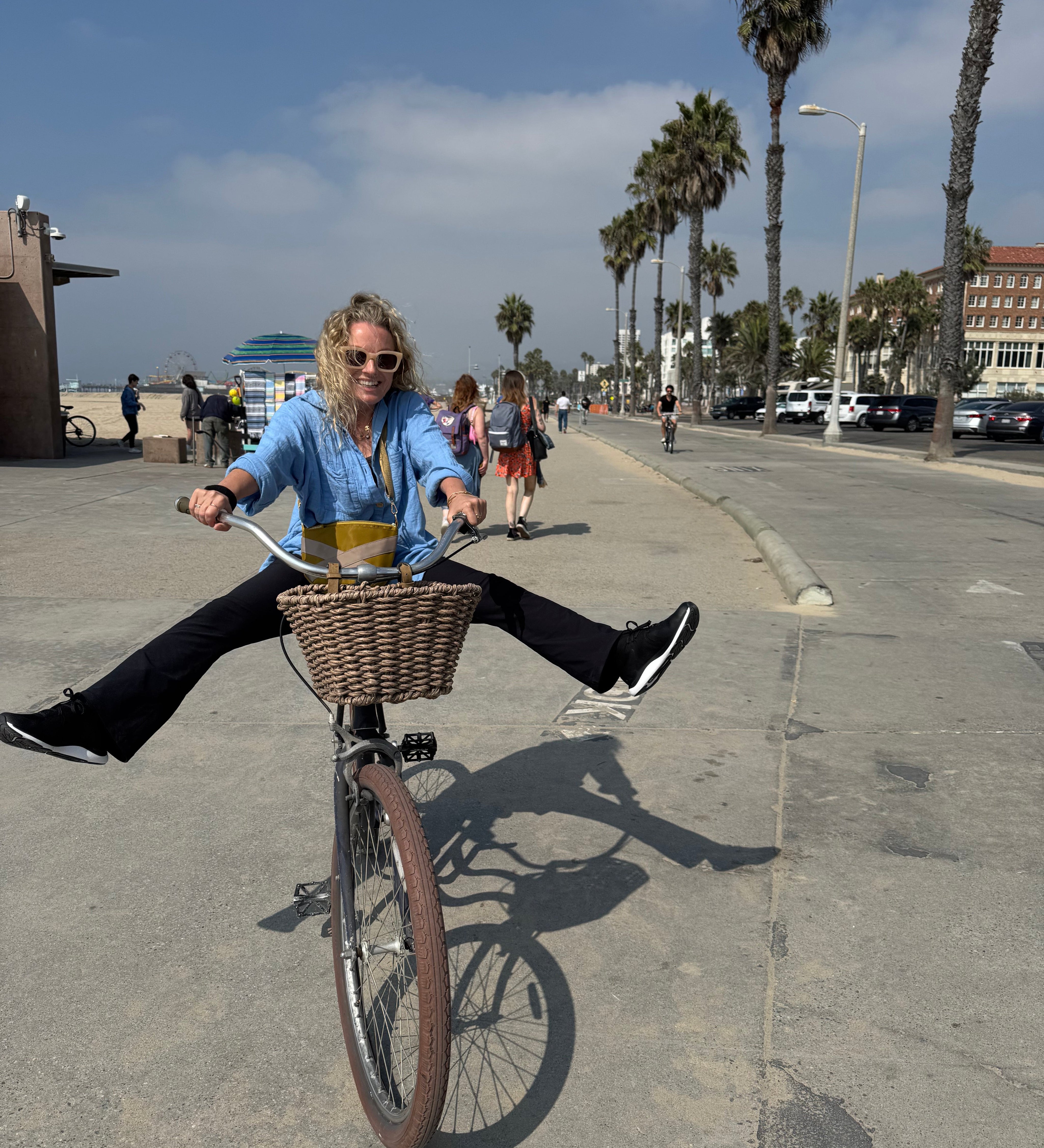 Lisa Walker, Co-Founder of Eir Women, riding a bike along the beachfront promenade in Los Angeles, smiling with legs outstretched on a sunny day.