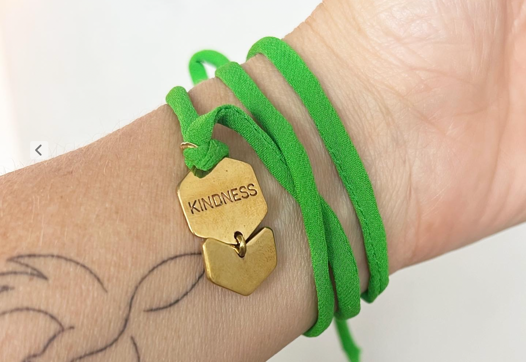 green string around wrist with metal charm that reads kindness, on a female wrist with small tattoo