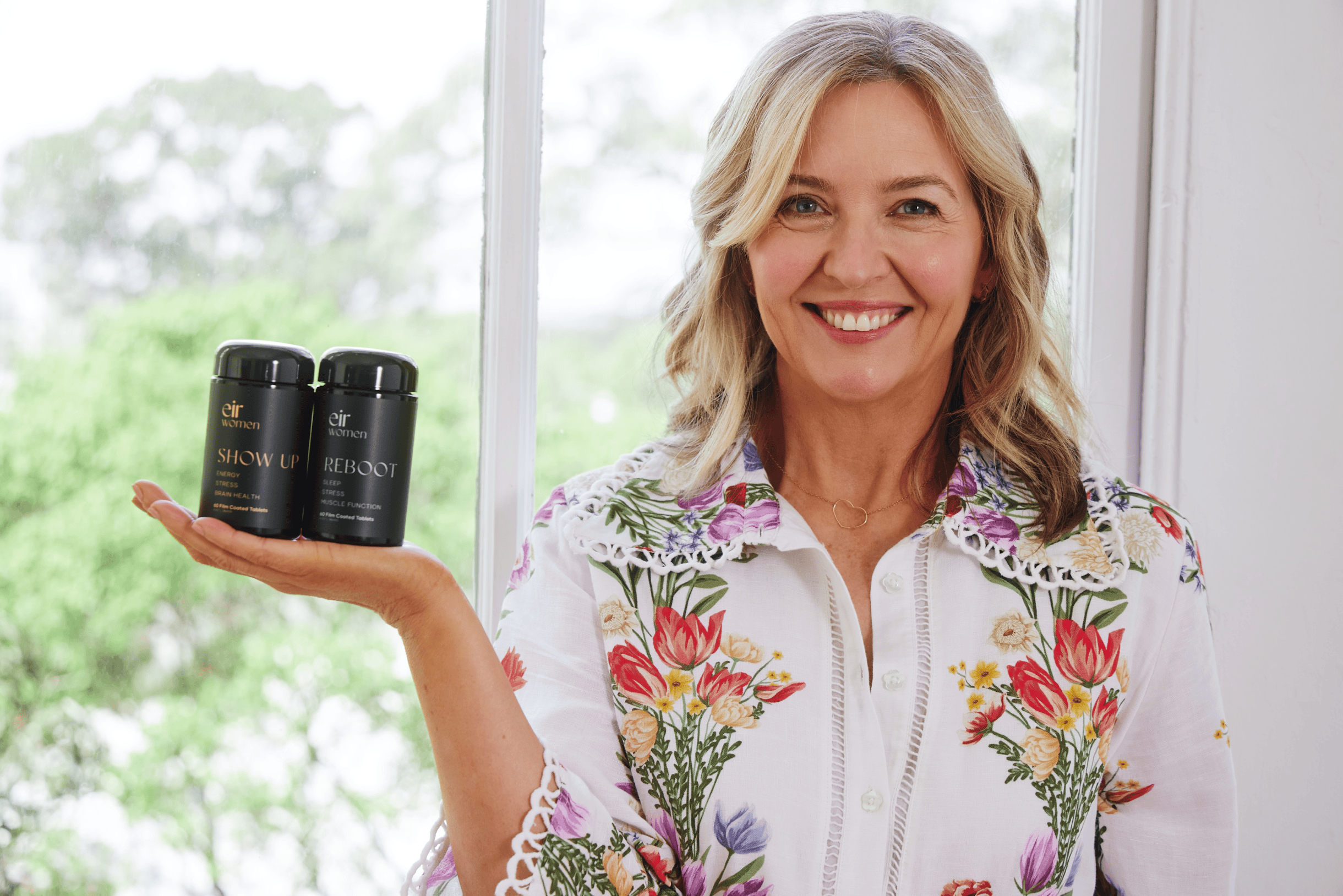 Kate Guaran, co-founder of Eir Women, smiling while holding two Eir supplement bottles—'Show Up' and 'Reboot'—with a bright window and greenery in the background.