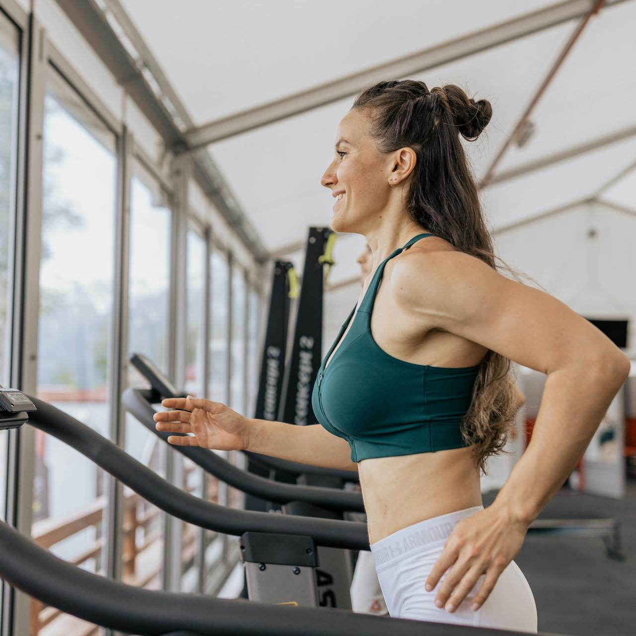Woman running on a treadmill in a gym, wearing activewear and smiling while exercising near large windows.