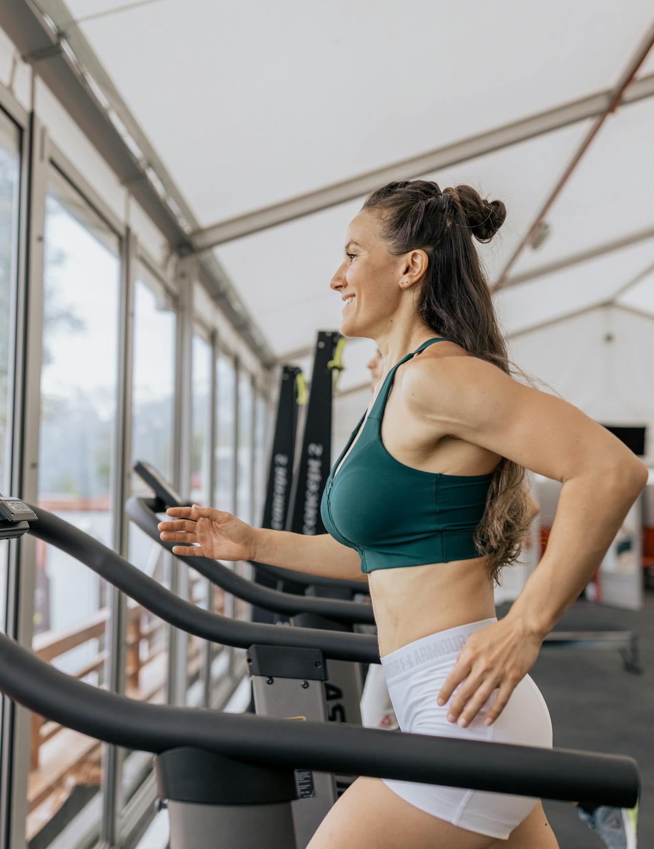 Woman running on a treadmill in a gym, wearing activewear and smiling while exercising near large windows.