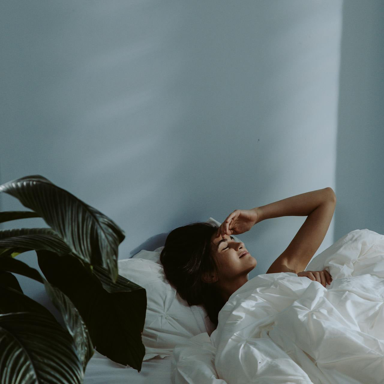 Woman lying in bed under white bedding, eyes closed with hand resting on her forehead, in a softly lit minimalist bedroom with a plant nearby.