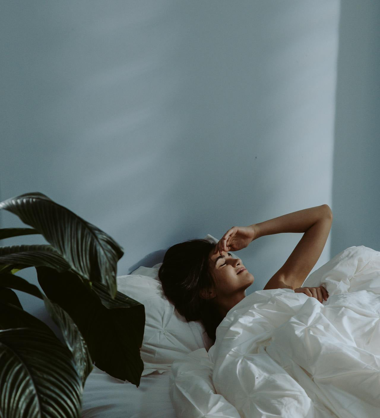 Woman lying in bed under white bedding, eyes closed with hand resting on her forehead, in a softly lit minimalist bedroom with a plant nearby.