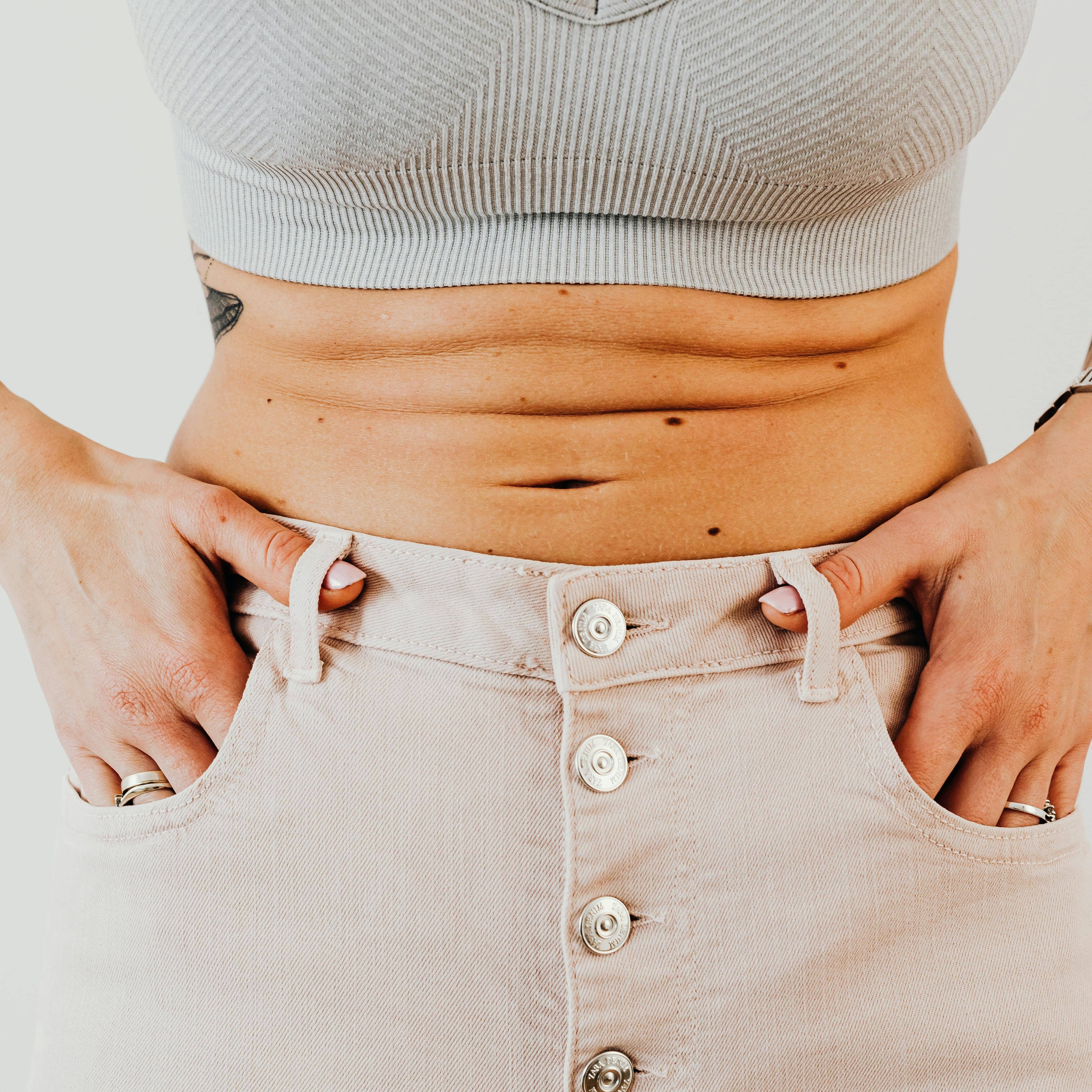 Close-up of a woman's midsection wearing high-waisted beige jeans and a ribbed crop top, showing natural belly folds and hands in pockets.