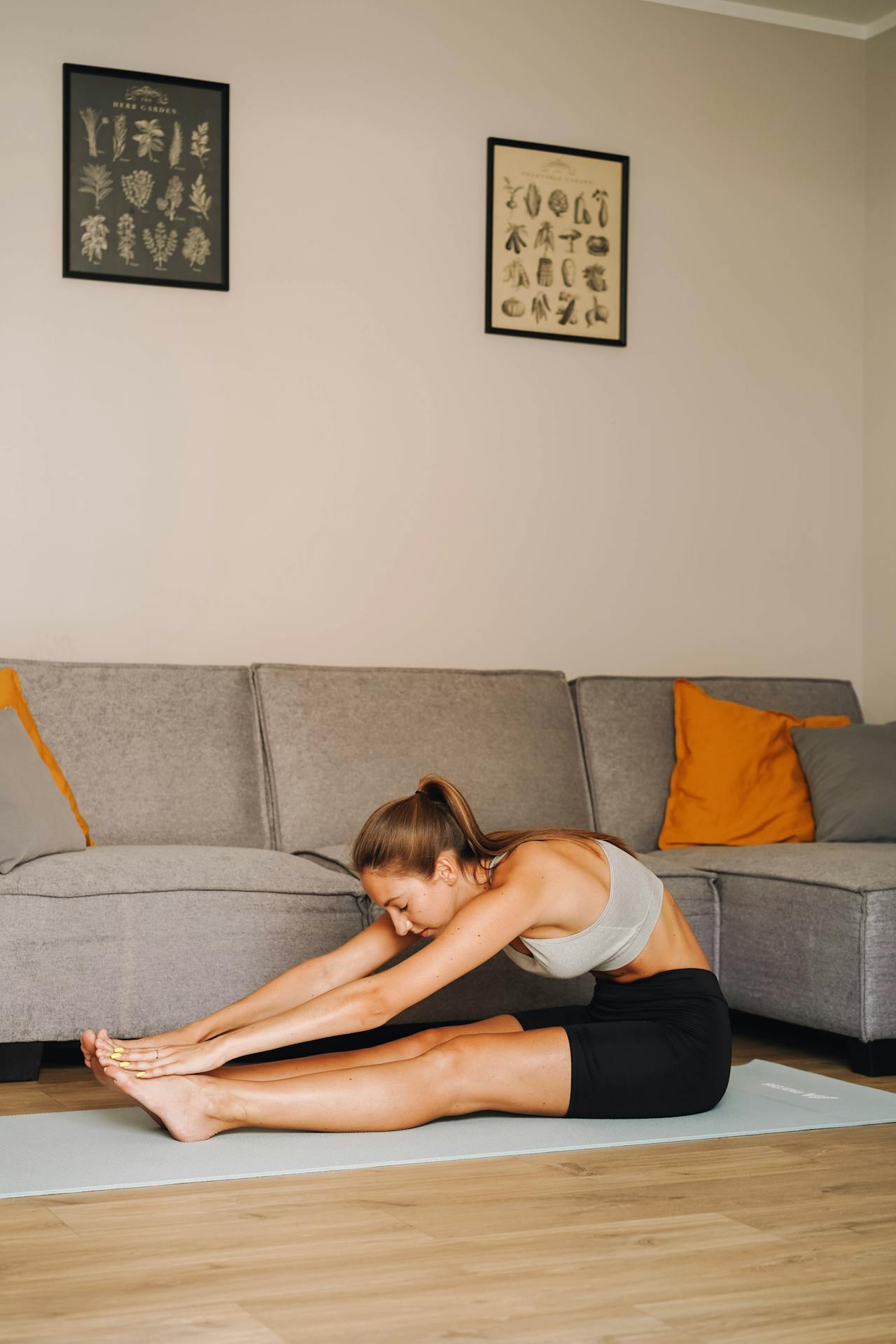 Woman stretching forward on a yoga mat in a living room, reaching for her toes during a seated stretch in front of a grey sofa.