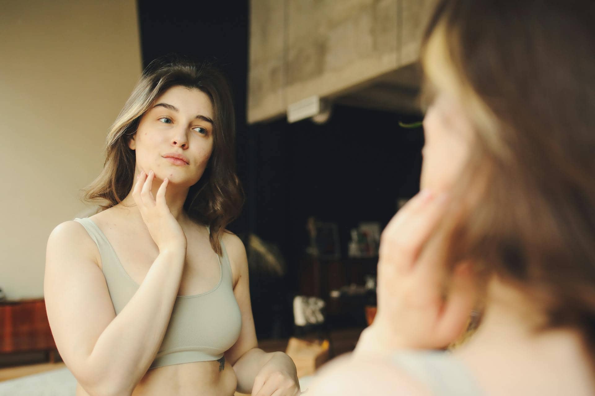 Woman with dark hair looking at reflection in mirror and touching face with fingers